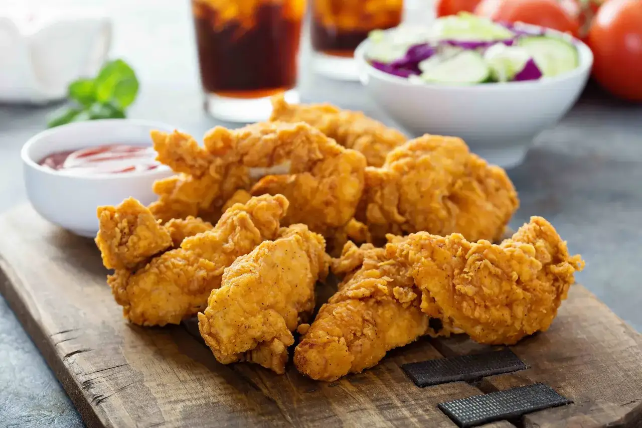 Croccanti strisce di pollo alla diavola su tagliere di legno, con salsa, insalata e bevande sullo sfondo.