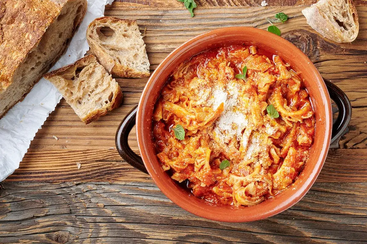 Trippa alla romana in terracotta, cosparsa di parmigiano, con pane casereccio a fette su un tavolo di legno.