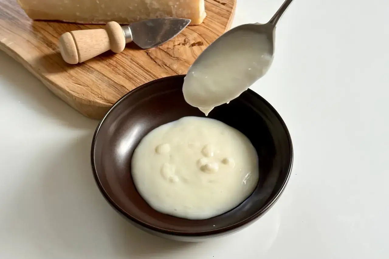 Una cucchiaiata di crema di parmigiano cade in una ciotola. Sul tagliere, un pezzo di formaggio e un coltellino.