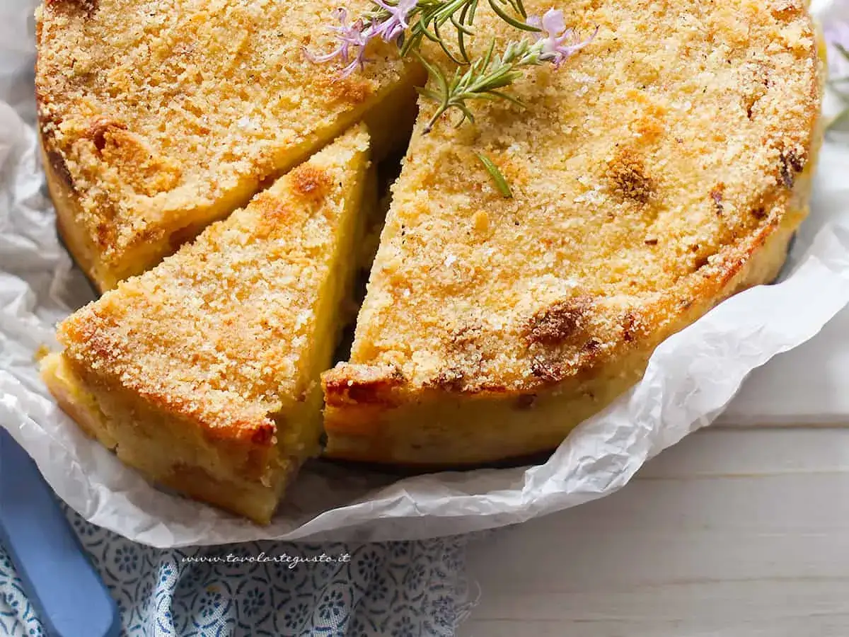 Gateau di patate Bimby dorato e croccante, guarnito con rosmarino. Una fetta &egrave; gi&agrave; stata tagliata, invitando ad assaggiarlo.