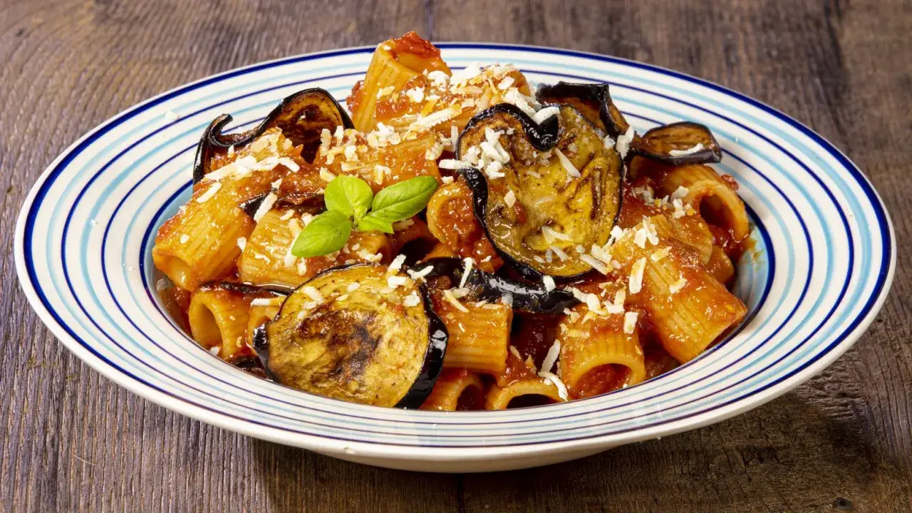 Piatto di pasta alla norma ricetta con melanzane fritte, pomodoro e ricotta salata grattugiata, guarnito con basilico fresco.
