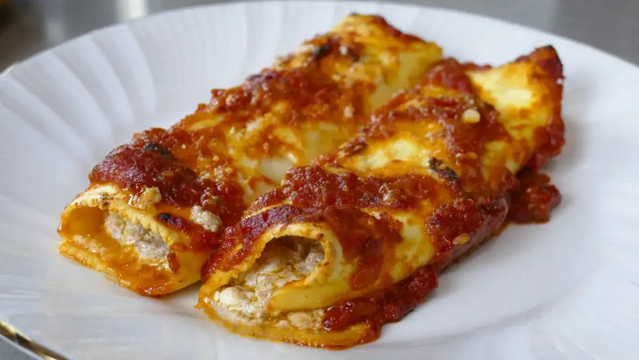 Due cannelloni al forno ripieni di carne e ricoperti di sugo rosso, serviti su un piatto bianco.
