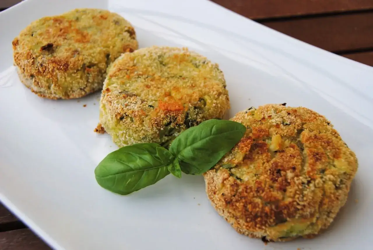 Tre polpette di zucchine e patate dorate e croccanti, guarnite con una foglia di basilico fresco su un piatto bianco.