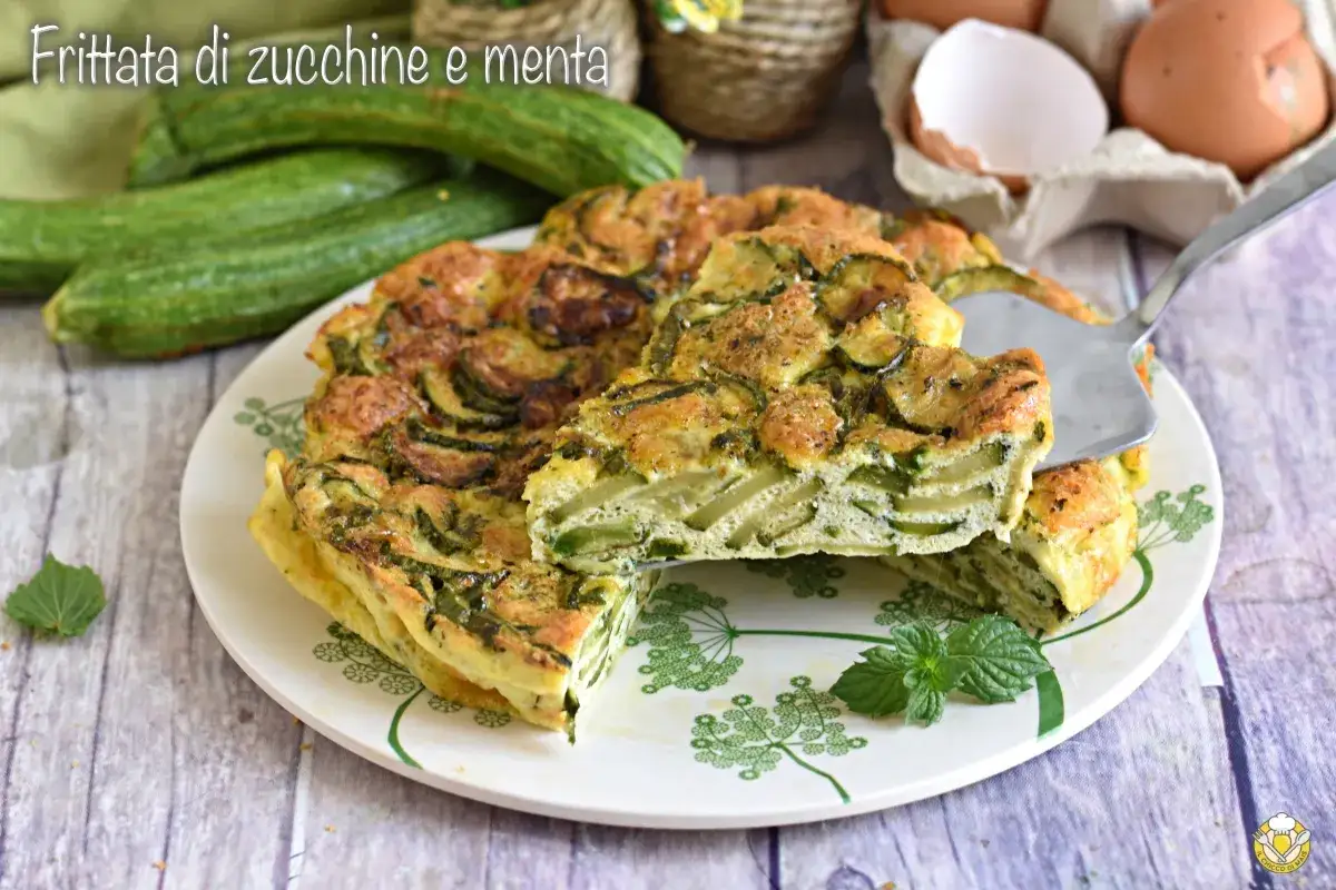 Una fetta di frittata di zucchine e menta, soffice e dorata, viene sollevata da un piatto decorato.