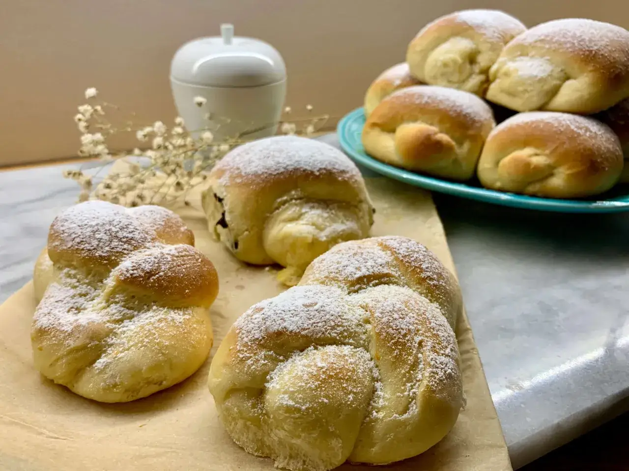 Deliziose brioche fatte in casa, spolverate di zucchero a velo, su carta da forno e un piatto turchese.