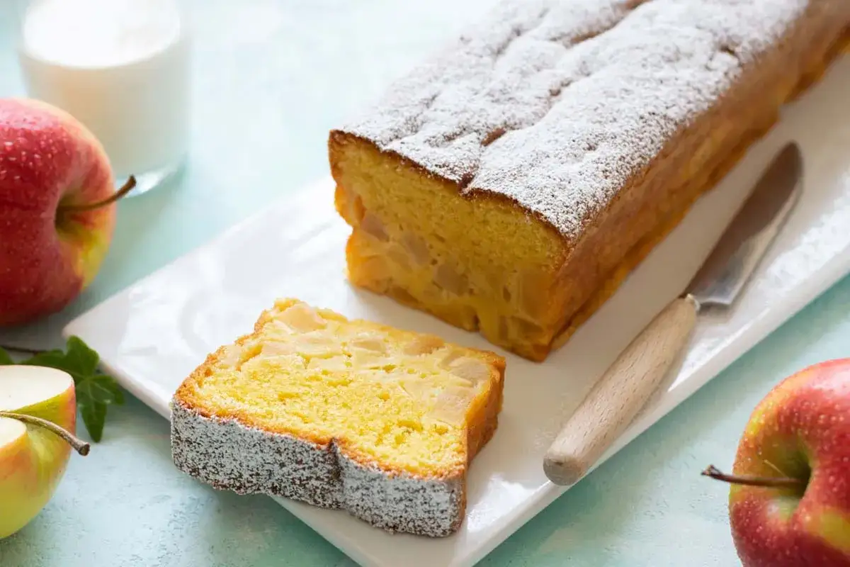 Un delizioso plumcake alle mele, cosparso di zucchero a velo, con una fetta gi&agrave; tagliata. Mele rosse e un bicchiere di latte completano la scena.