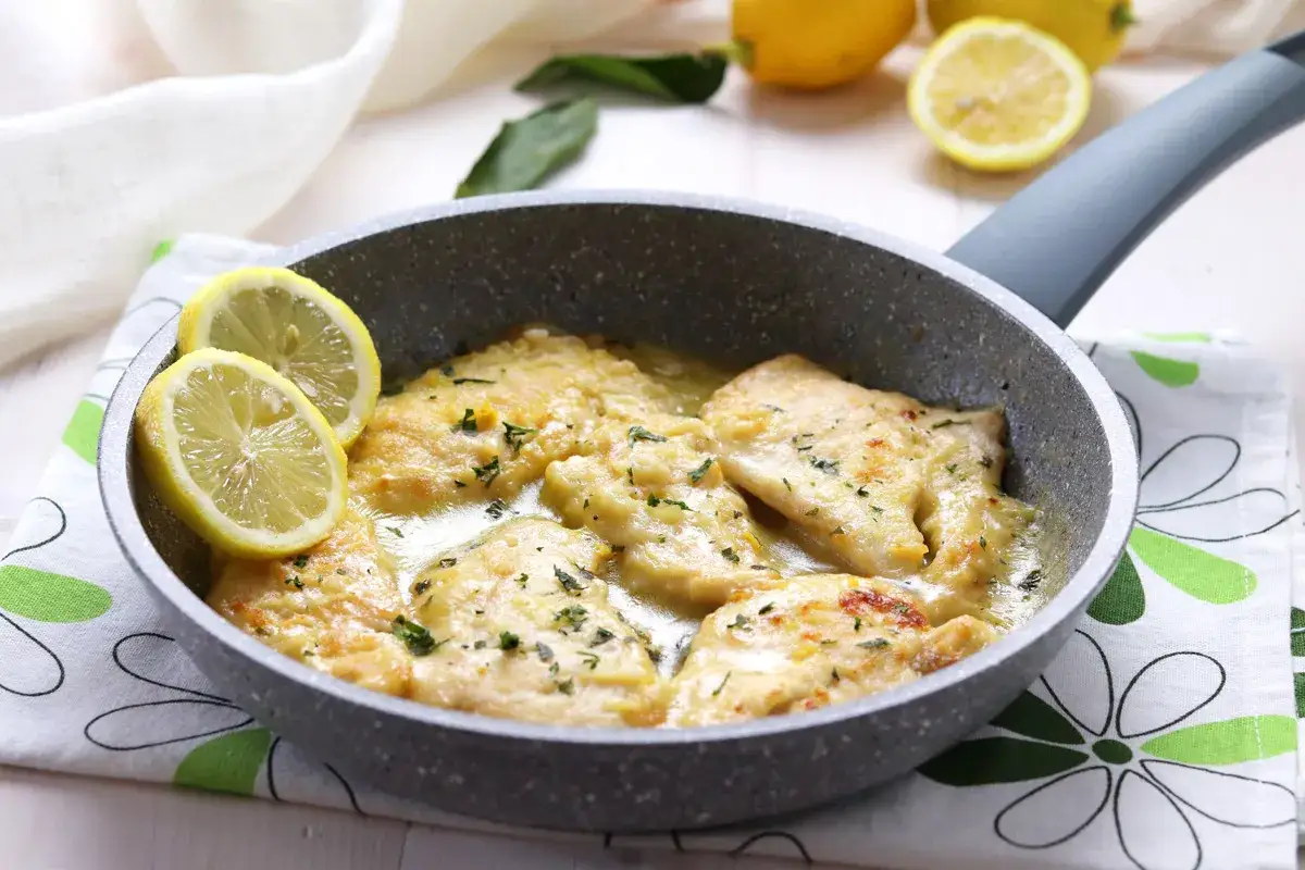 Scaloppine di pollo al limone in padella, guarnite con fette di limone fresco e prezzemolo tritato.