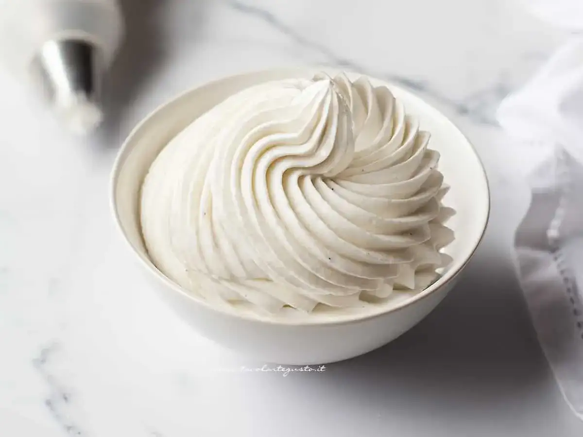 Una ciotola bianca piena di soffice crema al latte, con un beccuccio da pasticcere sfocato sullo sfondo.