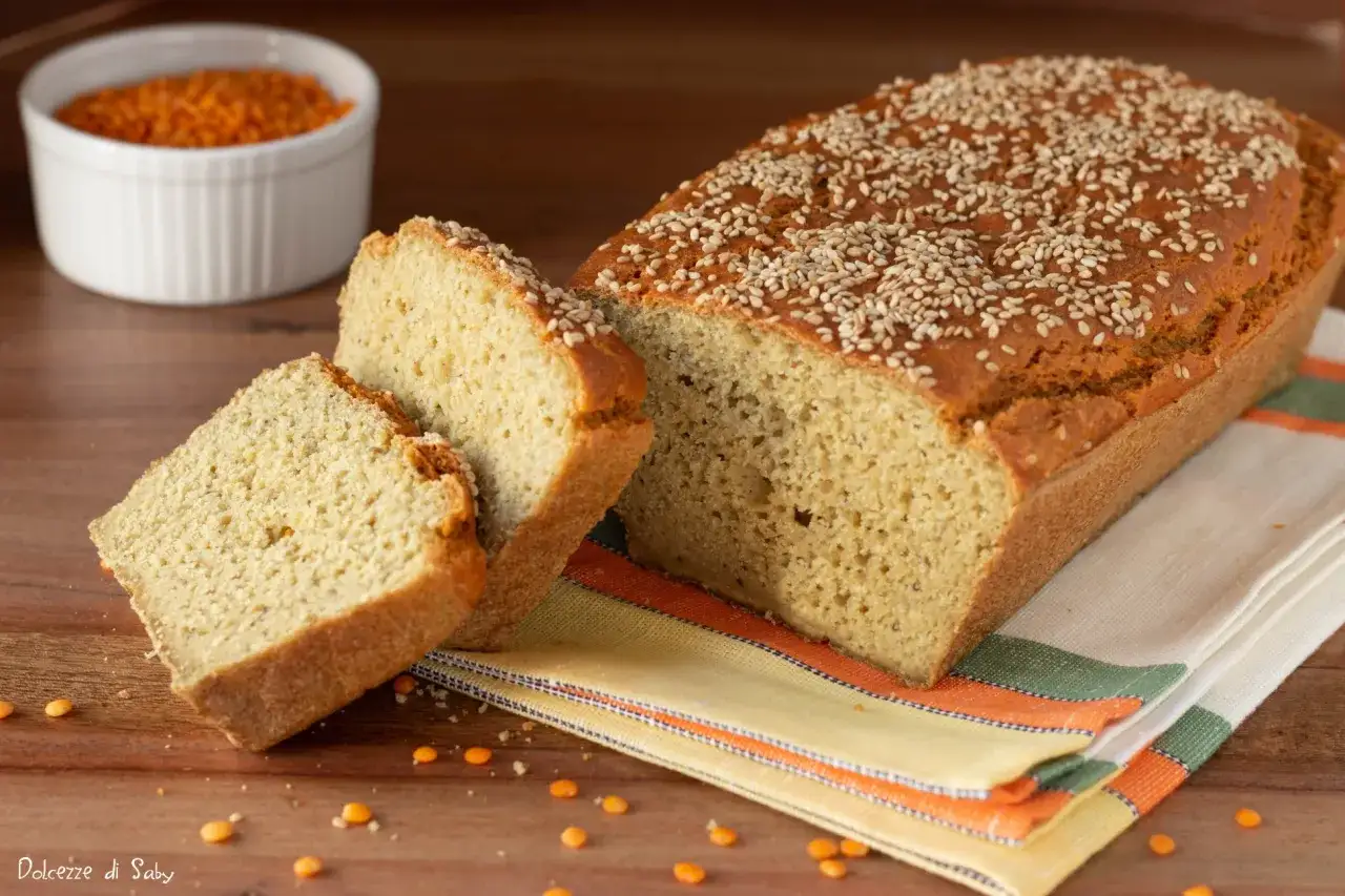 Pane di lenticchie dorato, cosparso di semi di sesamo, con due fette tagliate. Sullo sfondo, una ciotola di lenticchie rosse.