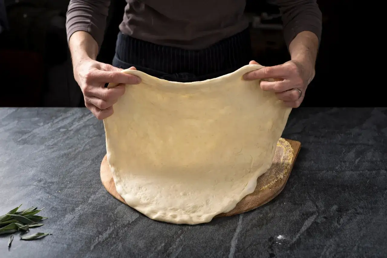 Mani che stendono un impasto soffice per pizza con lievito istantaneo, pronto per essere infornato.