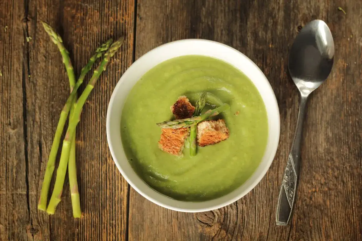 Una ciotola di cremosa crema di asparagi, guarnita con crostini e germogli freschi, accanto a un mazzo di asparagi su un tavolo di legno.