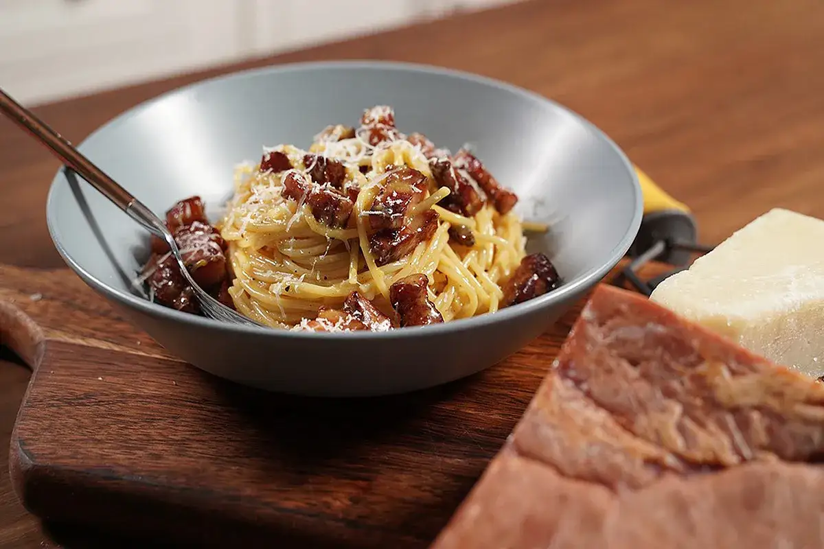 Un piatto di spaghetti alla gricia fumante, guarnito con guanciale croccante e pecorino. Accanto, pezzi di guanciale e formaggio.