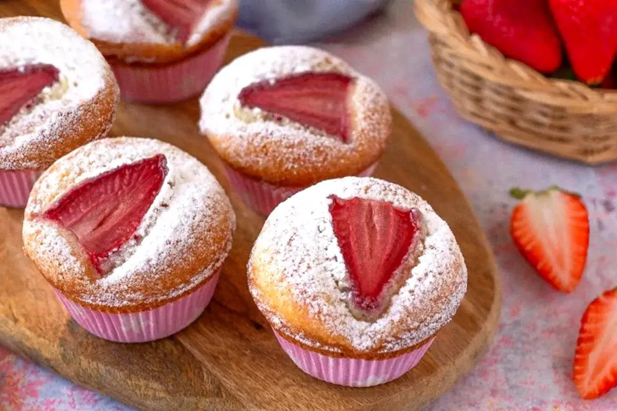 Muffin alle fragole spolverati di zucchero a velo, con fragole fresche sopra, su un tagliere di legno.