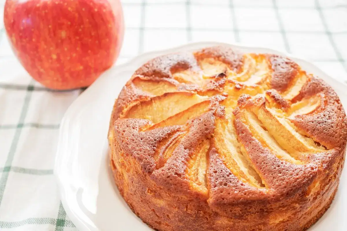 Torta di mele con yogurt, dorata e profumata, con spicchi di mela in superficie. Accanto, una mela rossa fresca.
