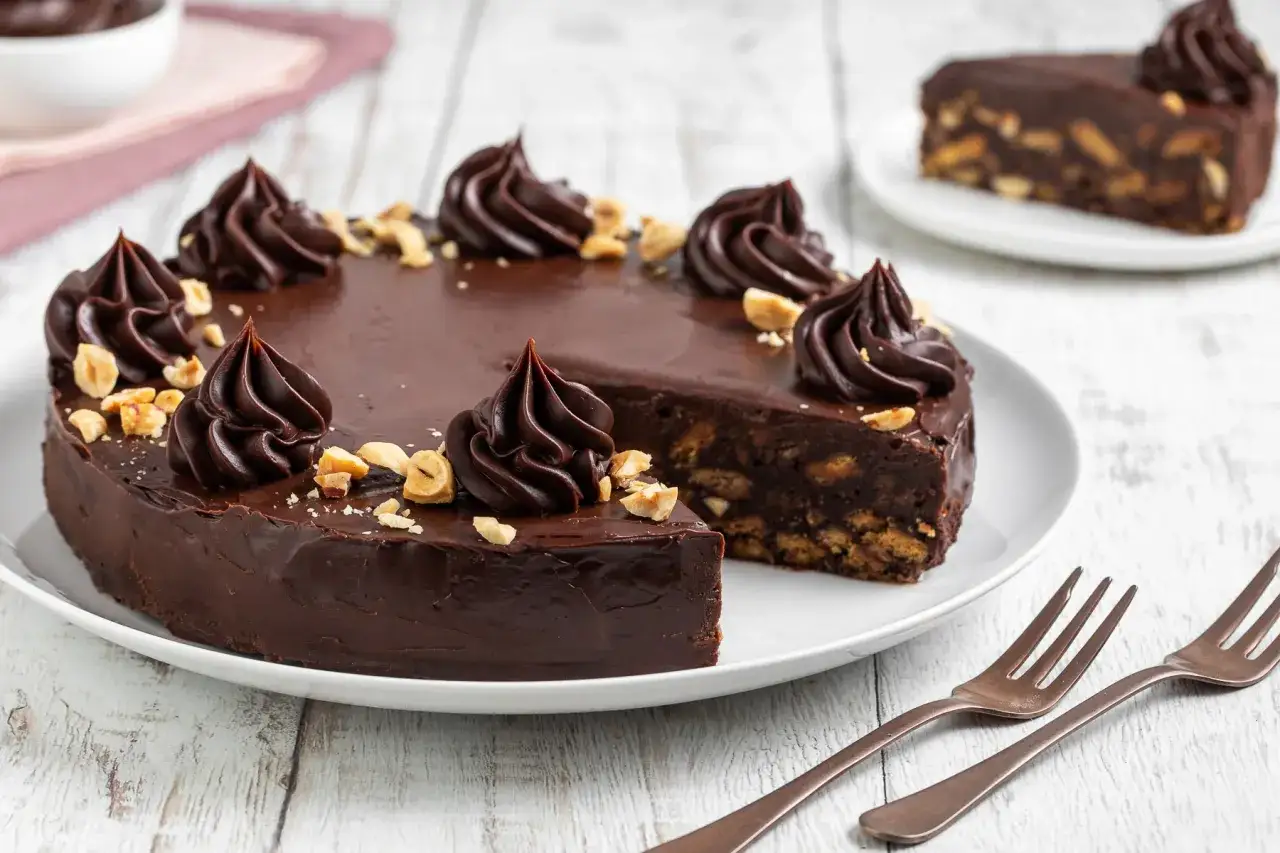 Un invitante dolce al cioccolato, ricoperto di glassa lucida e decorato con ciuffi di crema al cacao e nocciole tritate. Una fetta mostra l'interno ricco di biscotti.