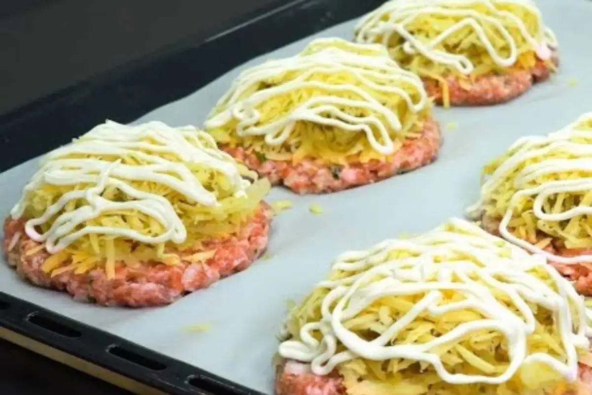 Hamburger al forno pronti per essere cotti, con carne macinata, formaggio grattugiato e un filo di maionese.