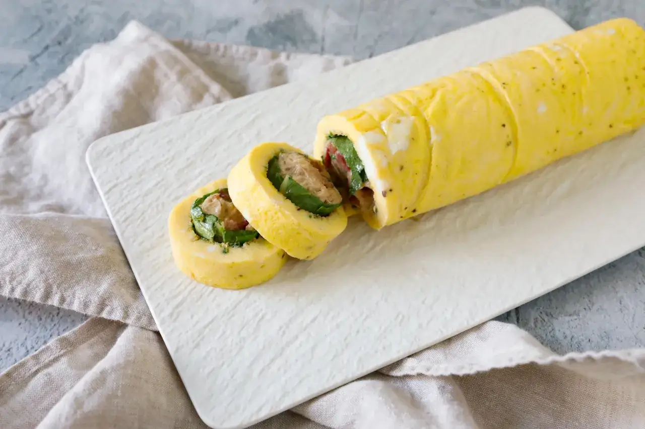 Rotolo di frittata giallo dorato, tagliato a fette, rivela un ripieno di verdure verdi e carne. Servito su un piatto bianco testurizzato.