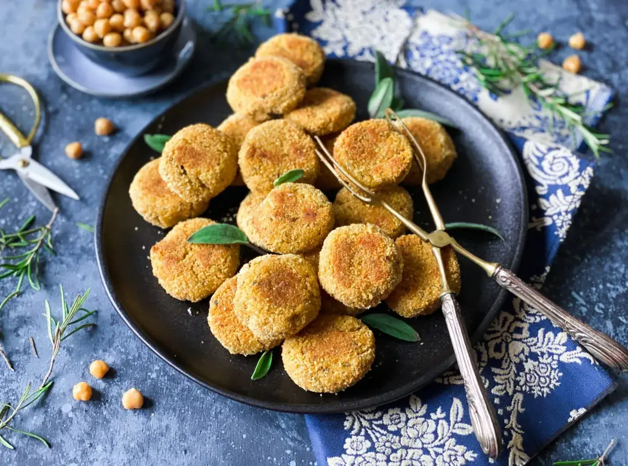 Piatto di polpette di ceci dorate, guarnite con foglie verdi, su un piatto nero. Vicino, forbici dorate e rametti di rosmarino.