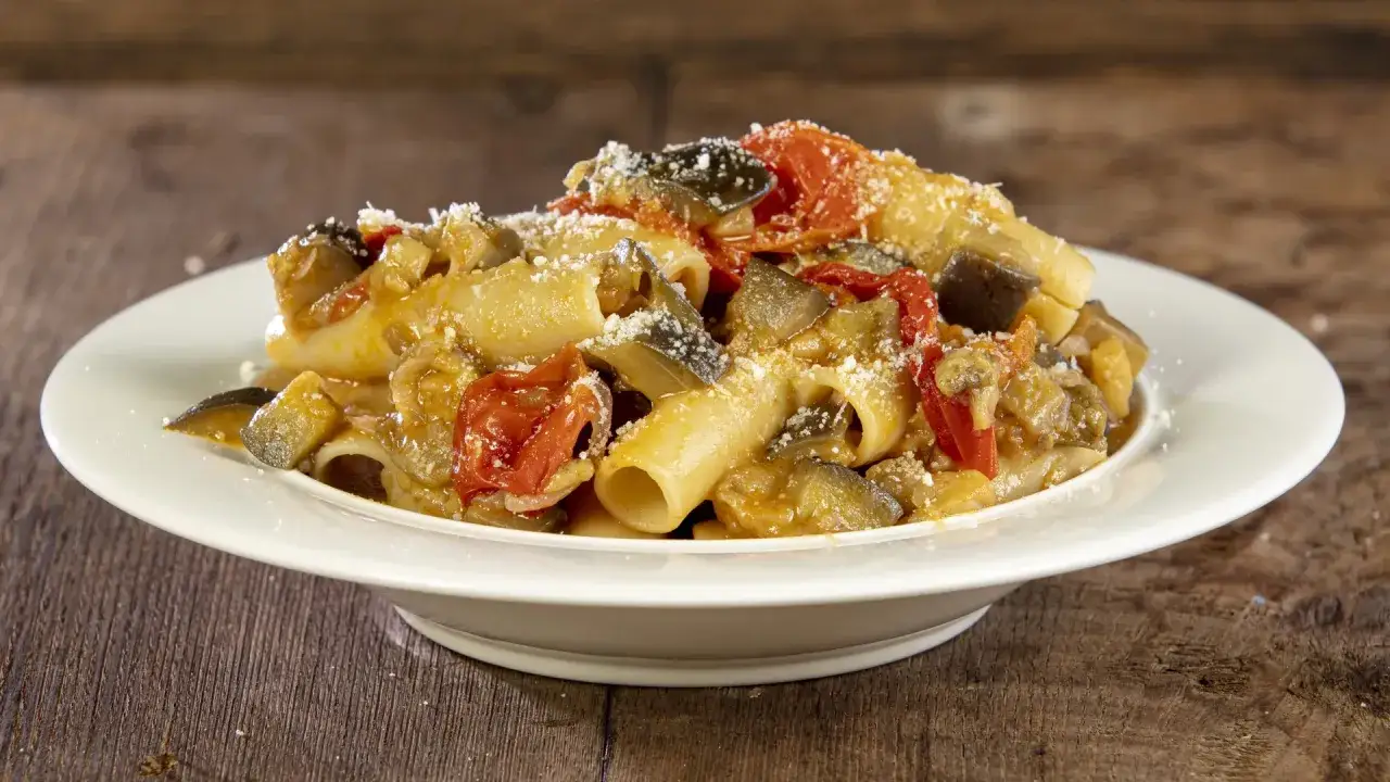 Un piatto di pasta con melanzane, pomodorini e formaggio grattugiato, servito su un piatto bianco.
