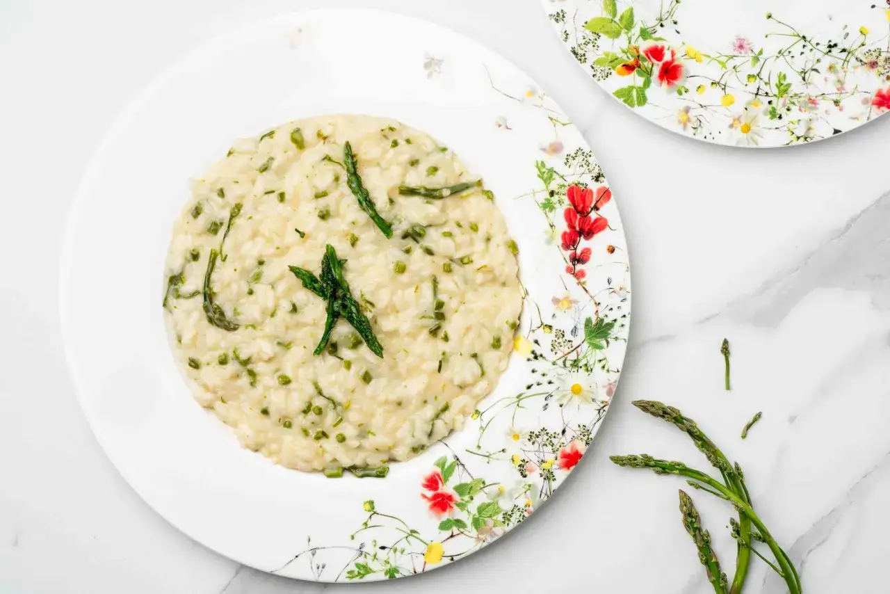 Un cremoso risotto con asparagi selvatici, guarnito con punte di asparagi freschi, servito su un piatto decorato con fiori primaverili.
