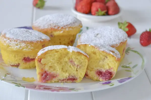 Morbidi muffin alle fragole, cosparsi di zucchero a velo, invitano a un dolce assaggio. Pezzi di fragola succosa si intravedono nell'impasto soffice.