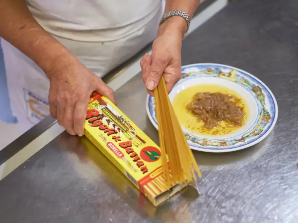 Mani che preparano i bigoli in salsa, con la pasta che esce dalla scatola e la salsa pronta nel piatto.